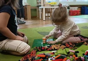 Dziewczynka siedzi na dywanie i buduje z klocków na zielonej podstawce.