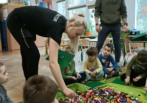 Pani Instruktor podaje dzieciom klocki. Wokół worka siedzą dzieci.