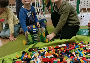 Dwóch chłopców buduje i uśmiecha się do zdjęcia. Na pierwszym planie widać leżące na podłodze klocki.