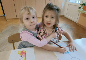 Dwie dziewczynki siedzą przy stoliku i przytulają się. Jedna z dziewczynek ma w ręce kredki.
