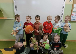 10 chłopców stoi na tle białej tablicy. W rękach trzymają pluszowe misie.