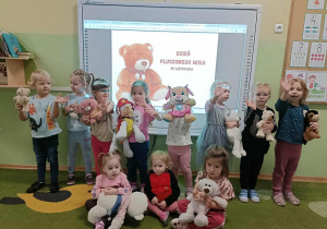 11 dziewczynek stoi na tle białej tablicy. Na tablicy jest napis "Dzień Pluszowego Misia". W rękach trzymają pluszowe misie.