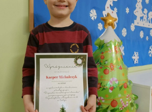 Chłopiec stoi z dyplomem w dłoni. Z prawej strony stoi choinka-balon. Z tyłu wisi tablica z niebieską dekoracją.