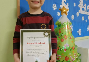 Chłopiec stoi z dyplomem w dłoni. Z prawej strony stoi choinka-balon. Z tyłu wisi tablica z niebieską dekoracją.