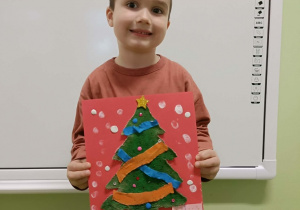 Chłopiec trzyma w dłoni pracę plastyczną .W tle biała tablica.