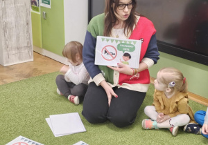 Pani Pedagog pokazuje dzieciom plansze. Obok niej siedzi dziewczynka w żółtej sukience oraz dziewczynka w białej bluzce. Na zielonym dywanie leżą plansze. W tle wisi czarny monitor.