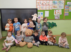 Grupa dzieci stoi i siedzi na zielonym dywanie. Dzieci w dłoniach trzymają pluszowe misie.