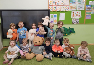 Grupa dzieci stoi i siedzi na zielonym dywanie. Dzieci w dłoniach trzymają pluszowe misie.