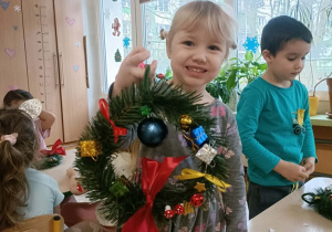 Dziewczynka w szarej sukience trzyma w dłoni skończony wianek.