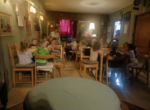 Grupa dzieci siedzi przy stolikach. W tle widać czerwoną zasłonę w oknie.