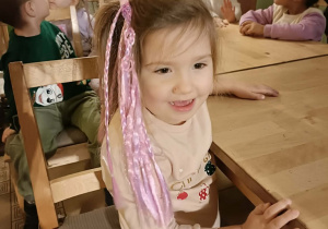 Dziewczynka w różowych warkoczykach siedzi przy stoliku i uśmiecha się do zdjęcia.