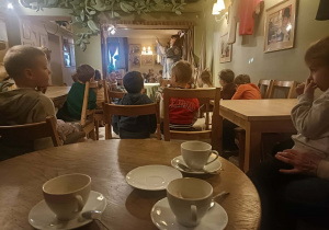 Dzieci oglądają spektakl, siedząc przy stolikach.