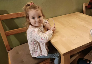 Dziewczynka w jasnej bluzce siedzi na krzesełku i uśmiecha się do zdjęcia.