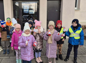 Grupa dzieci stoi przez budynkiem Szkoły Podstawowej. W rękach trzymają wykonane prace plastyczne.