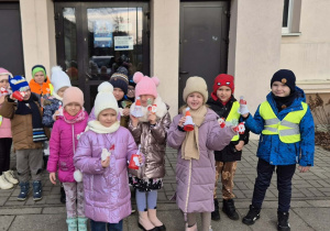 Grupa dzieci stoi przez budynkiem Szkoły Podstawowej. W rękach trzymają wykonane prace plastyczne.