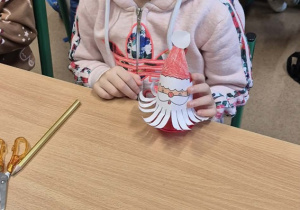 Dziewczynka w różowej bluzie siedzi przy stoliku i pokazuje wykonanego Mikołaja.