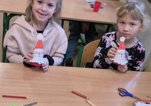 Dwie dziewczynki siedzą przy stoliku, uśmiechają się do zdjęcia i pokazują wykonane Mikołaje. W tle siedzi dwóch chłopców.