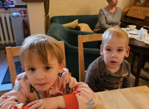 Dwóch chłopców siedzi przy drewnianym stole. Z tyłu widać szarą kanapę i szary fotel.