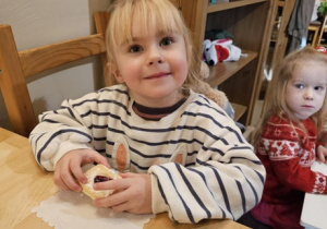 Dziewczynka w czarno-białej bluzie siedzi przy drewnianym stole. W tle widać dziewczynkę w czerwonym sweterku.