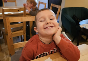 Chłopiec uśmiecha się do zdjęcia. Na stole leży ciastko. W tle widać dzieci siedzące przy innych stolikach.