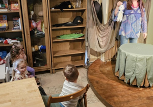 Po prawej stronie scena, na której występuje Pani w niebieskim stroju. Po lewo przy stoliku siedzą dzieci.