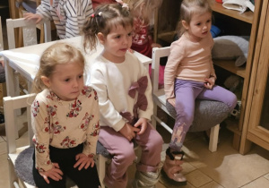 Trzy dziewczynki siedzą na krzesełkach i patrzą przed siebie, W tle widać dzieci siedzące przy innych stolikach.