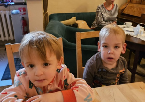 Dwóch chłopców siedzi przy drewnianym stole. Z tyłu widać szarą kanapę i szary fotel.