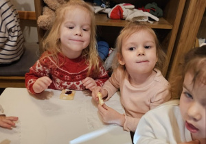 Trzy dziewczynki siedzą przy stolikach i jedzą ciastka. W tle widać drewniany regał. Na jednej z półek widoczny jest napis "czapki".