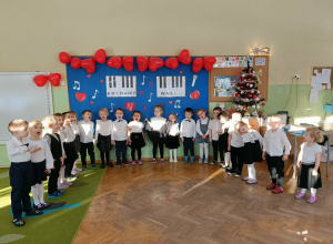 Dzieci stoją na tle dekoracji. Na dekoracji jest napis "Kochamy was". Na górze tablicy wiszą balony w kształcie serc. Po prawej stronie stoi choinka.