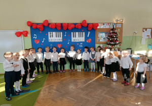 Dzieci stoją na tle dekoracji. Na dekoracji jest napis "Kochamy was". Na górze tablicy wiszą balony w kształcie serc. Po prawej stronie stoi choinka.