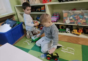 Chłopiec w bluzce w paski trzyma w dłoni zielone auto. Chłopiec w szarej bluzce zdejmuje auto z regału.