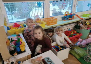 Cztery dziewczynki bawią się w kąciku kuchennym. Na drugim planie parapet, na którym stoją zółte pojemniki z zabawkami.