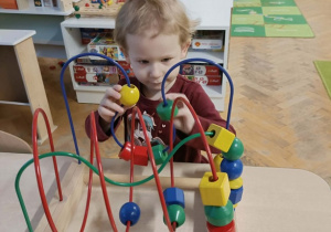 Dziewczynka w krótkich włosach siedzi przy stoliku i bawi się drewnianą, kolorową przeplatanką.