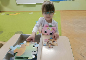 Dziewczynka w bluzce w kotki siedzi przy stoliku i układa puzzle z Psim Patrolem.