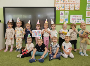 Grupa dzieci stoi na tle tablicy multimedialnej. Na głowach dzieci mają czapki.