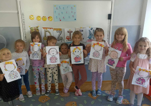 9 dziewczynek stoi na tle białej tablicy z wykonaną pracą plastyczną.