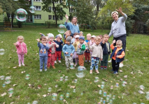 Dzieci stoją na trawie wraz z Paniami i uśmiechają się do zdjęcia.