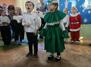 Chłopiec w białej koszuli i dziewczynka przebrana za choinkę recytują wiersz. W tle stoją dzieci.