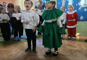 Chłopiec w białej koszuli i dziewczynka przebrana za choinkę recytują wiersz. W tle stoją dzieci.