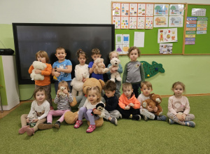 Grupa dzieci stoi i siedzi na zielonym dywanie. Dzieci w dłoniach trzymają pluszowe misie.