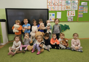 Grupa dzieci stoi i siedzi na zielonym dywanie. Dzieci w dłoniach trzymają pluszowe misie.