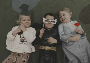 Dwie dziewczynki i chłopiec leżą na dywanie z rekwizytami do fotobudki.