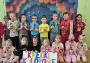 Dzieci stoją na tle kolorowej dekoracji. W dłoniach trzymają papierowe kredki.