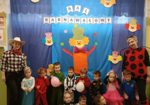 Grupa dzieci wraz z Paniami stoi na tle niebieskiej dekoracji. Na płótnie napis "Bal karnawałowy" oraz klauny.