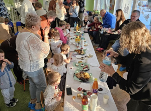 Na stole biały obrus. Wokół stołu siedzą dzieci. Dziadkowie stoją obok dzieci.