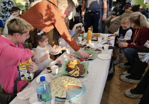 Na stole biały obrus. Wokół stołu siedzą dzieci. Dziadkowie stoją obok dzieci.