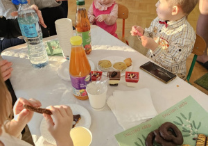 Przy stole siedzi chłopiec i dziewczynka. Dziewczynka ma różową sukienkę i opaskę na głowie.