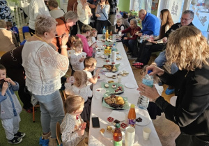 Na stole biały obrus. Wokół stołu siedzą dzieci. Dziadkowie stoją obok dzieci.