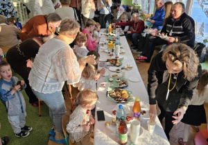 Na stole biały obrus. Wokół stołu siedzą dzieci. Dziadkowie stoją obok dzieci.