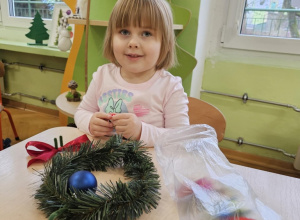 Dziewczynka uśmiecha się do zdjęcia. Na stole leży wianek oraz ozdoby w woreczku.